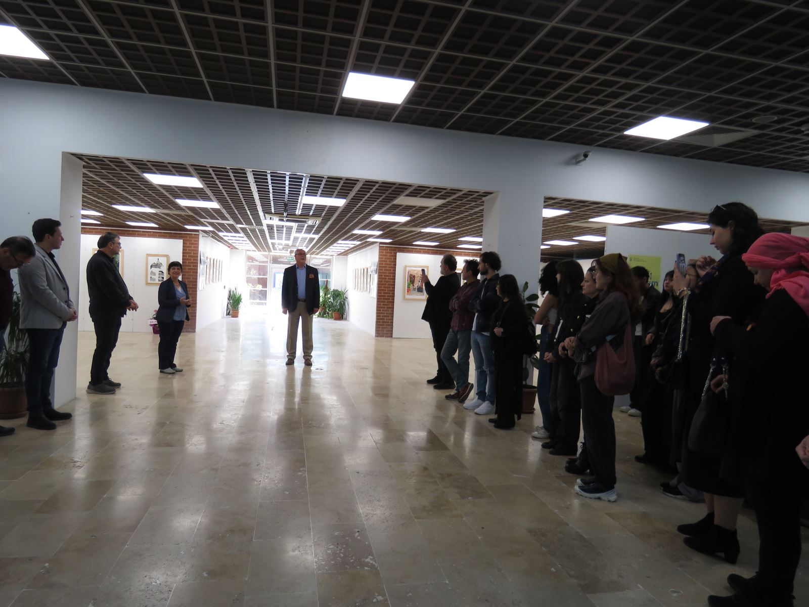 Sergi açılışında konuşmalar yapılıyor. Sergi salonunda öğrenciler ve akademisyenler görünmektedir.